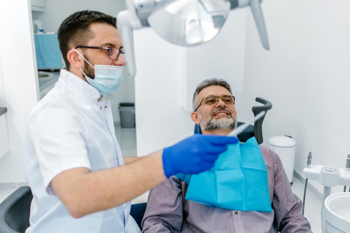 A middle aged man sits in a dentist’s chair, speaking to his dentist, who is standing beside him.