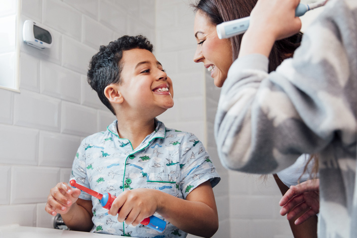 Children are brushing their teeth in the bathroom at home. The mother is checking the little boy's mouth to make sure he has brushed properly.