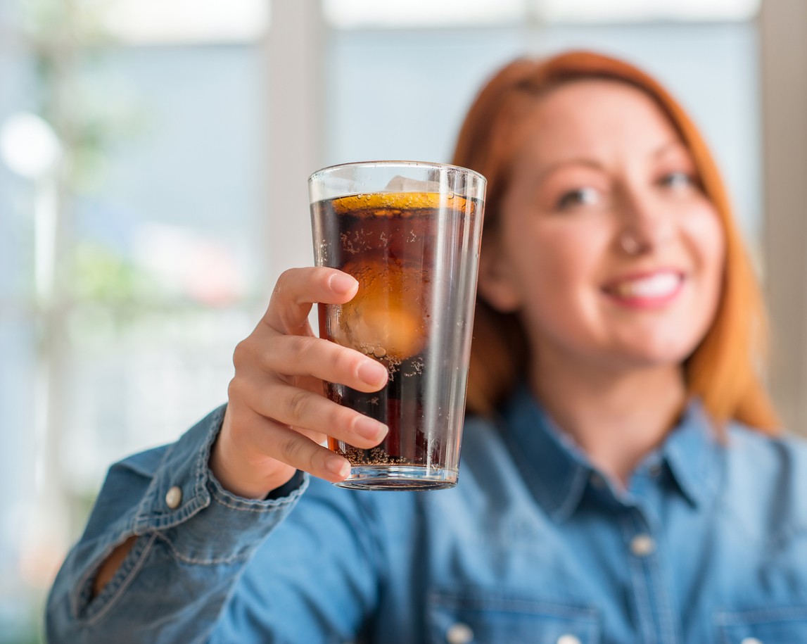 Redhead woman holding soda refreshment with a happy face standing and smiling with a confident smile showing teeth